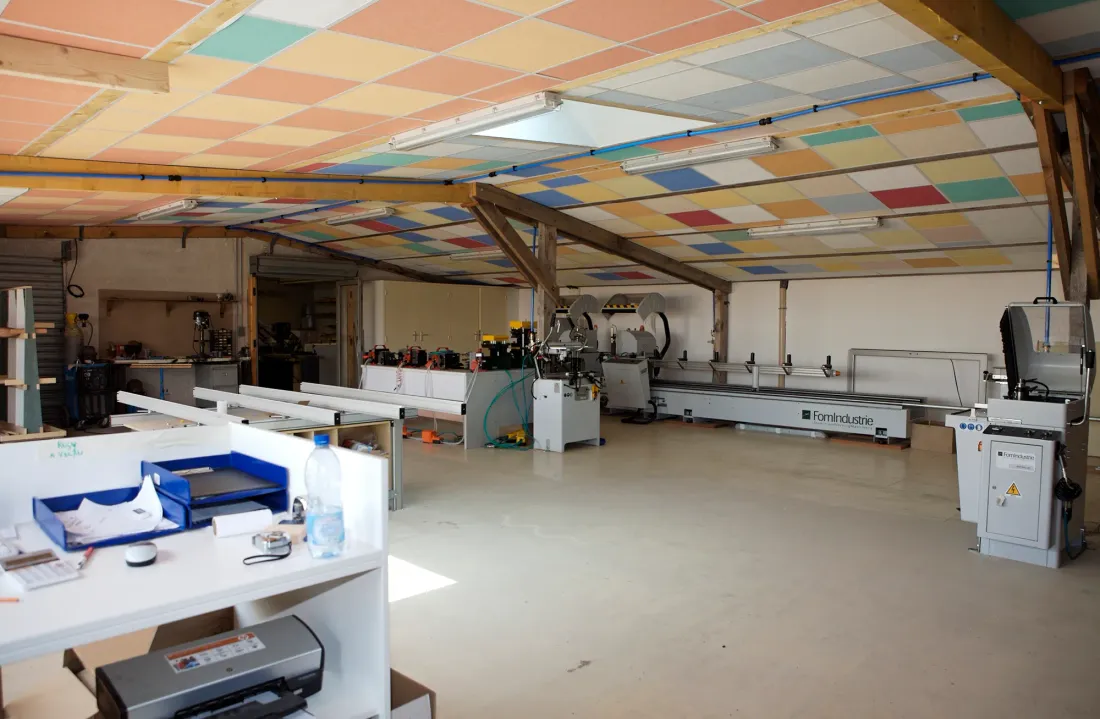 Atelier de menuiserie : plafond en panneaux colorés, plaqueuse de chants et longs établis sous charpente en bois.