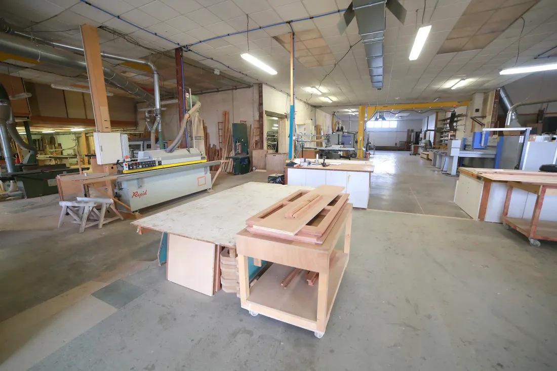 Atelier de menuiserie : établi mobile, panneaux de bois, machines et système d'aspiration sous charpente métallique.