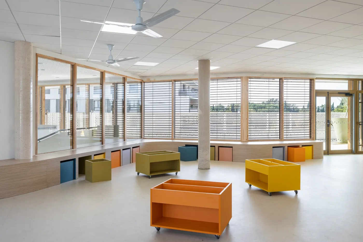Grande salle d'un groupe scolaire, banquette cintrée en bois le long de la baie